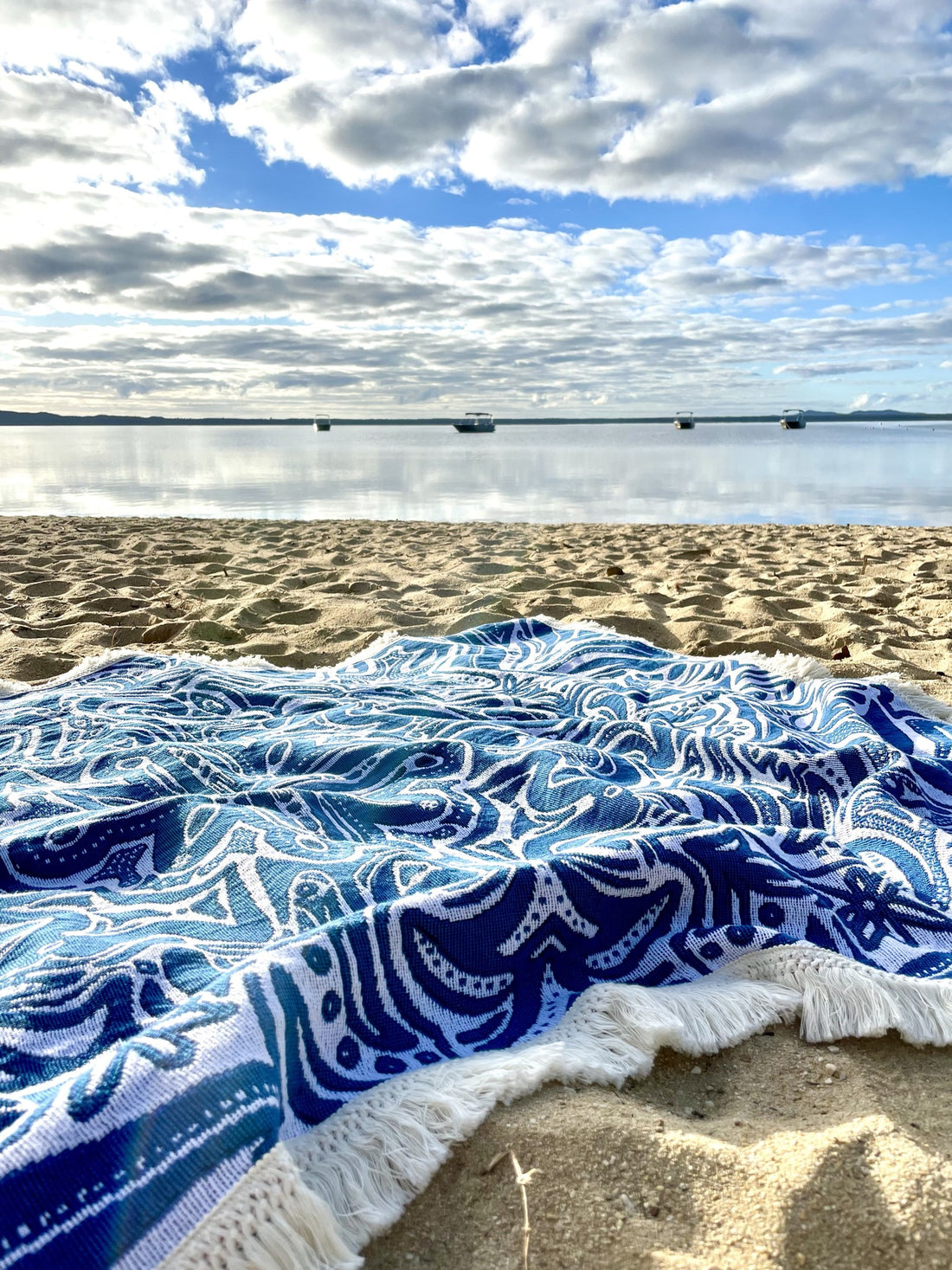Blue Skies beach blanket or throw rug laying on sand overlooking still water. Designed in Australia. 