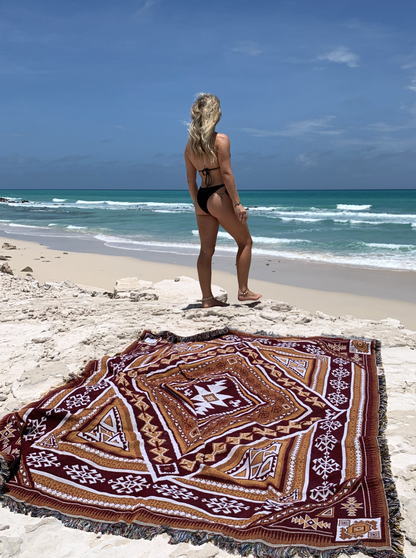 Desert Night throw rug and picnic blanket laying on beach, crystal blue water in background. Shades of brown, boho rug, must-have van life accessory. Designed in Australia for adventure.