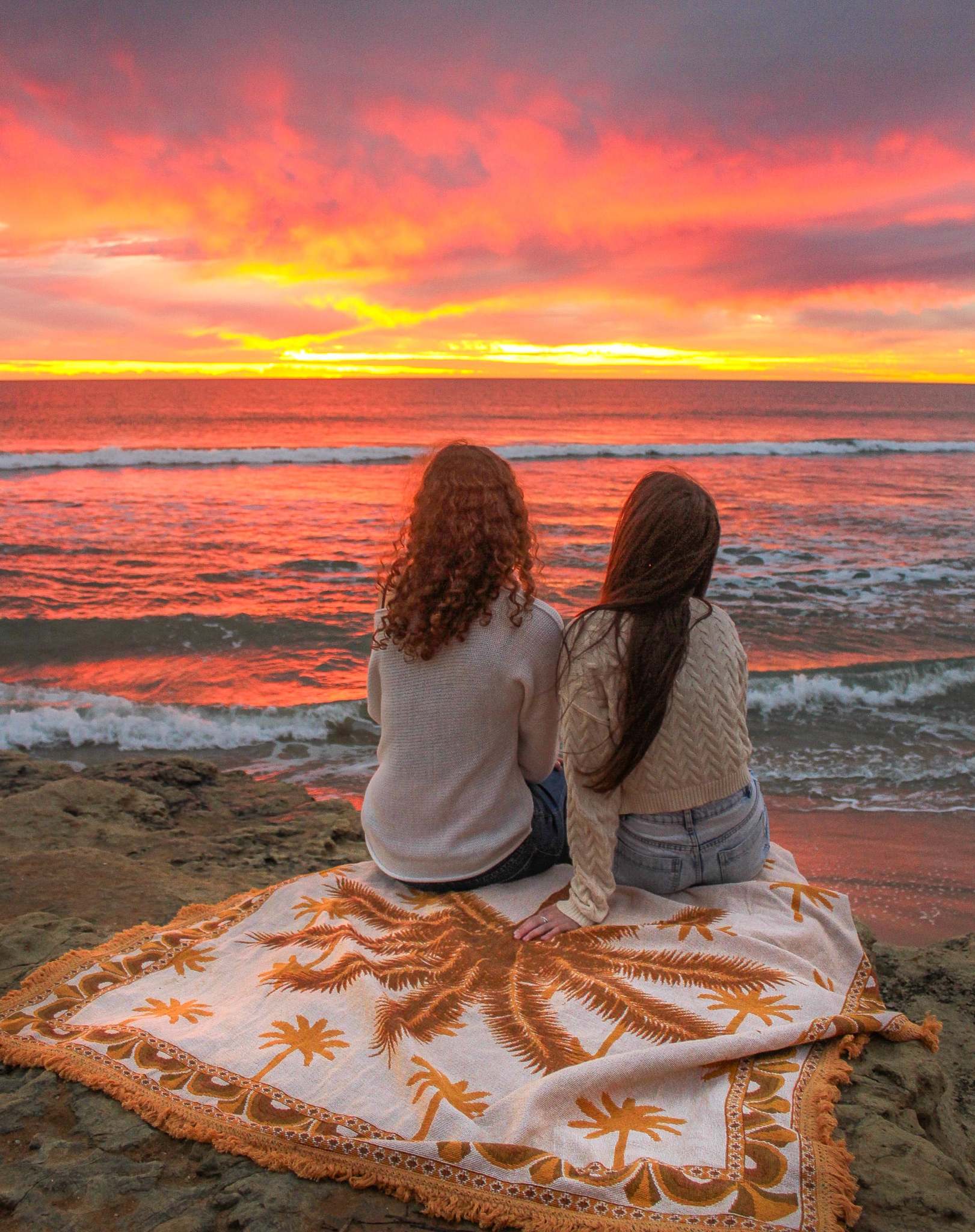 Endless Summer throw rug on rocks by the ocean, two girls sit in warm clothes looking out to orange skies as the sun sets