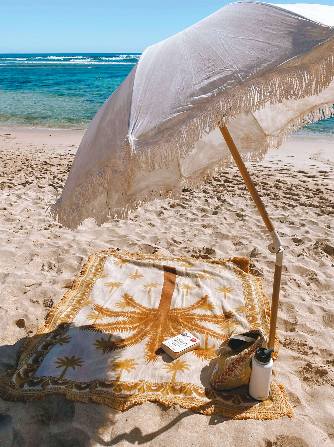 Endless Summer Palm Throw Blanket on the beach under a cream umbrella, clear blue ocean in the background. Ready for self-care - book and waterbottle on hand
