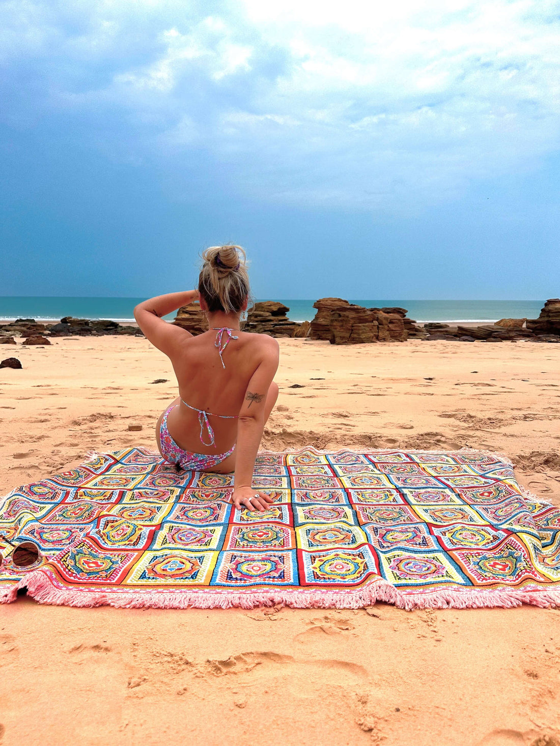 Forever Young throw rug on the sand overlooking a storm rolling in over the ocean. Young woman sits on throw beach blanket looking out.