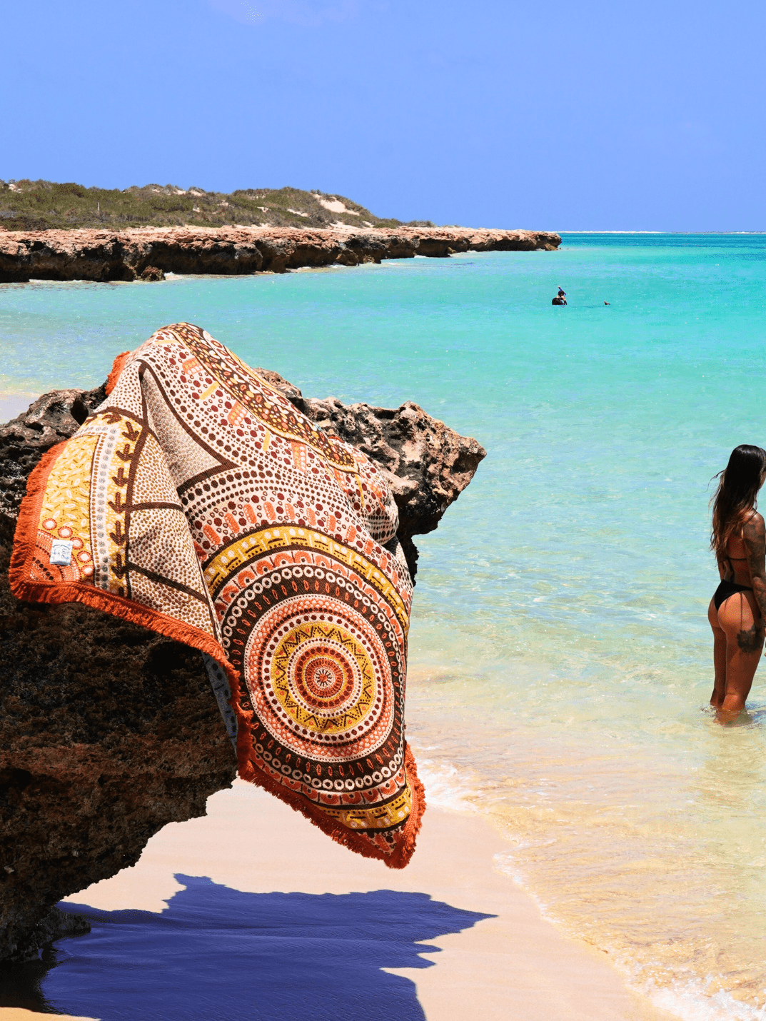 Stomping Ground throw rug design, Indigenous Aboriginal art beach blanket in orange and brown tones hanging over rocks on the beach, crystal blue ocean in background. Western Australia